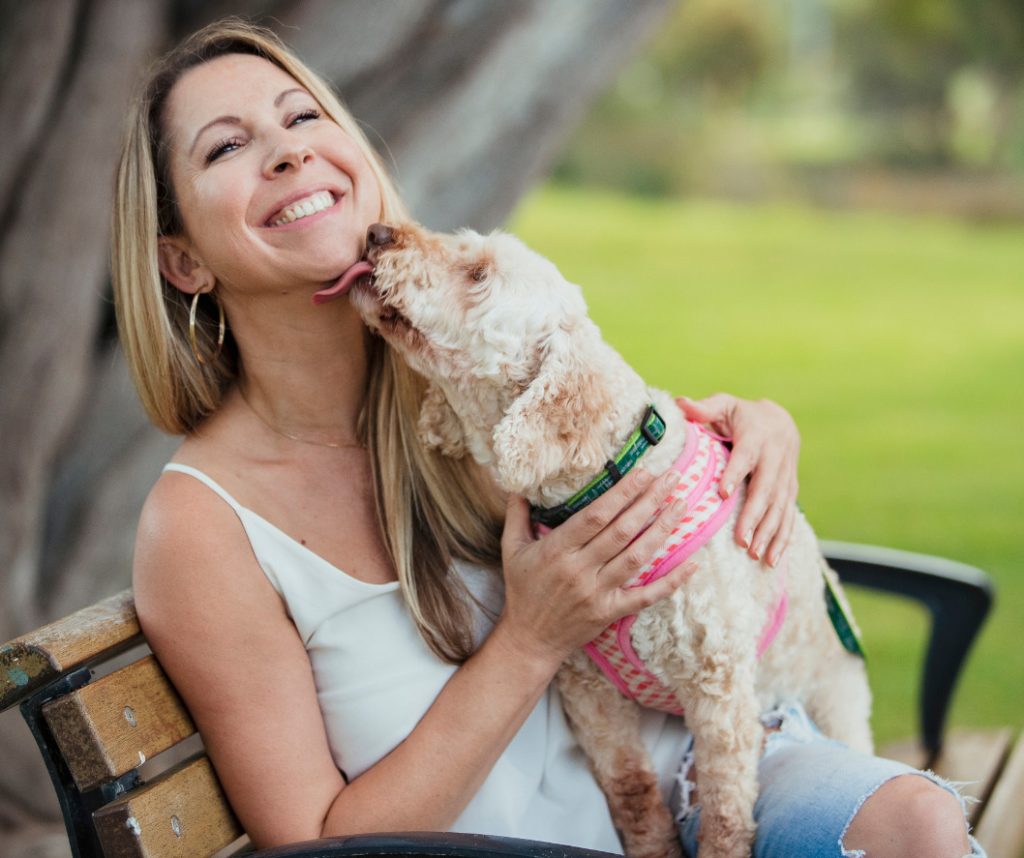 Mujer sonriente sentada en un banco del parque mientras su perro la lame cariñosamente en la cara. La imagen refleja la alegría y el afecto mutuo en su relación, capturando un momento espontáneo y feliz entre ambos.